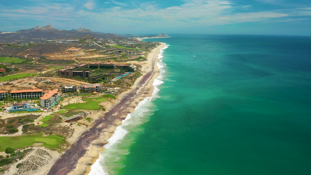 Aerial view of the San Jose del Cabo coastline at the start of the clip, with turquoise water on the right, sandy beach and dark shoreline bands below, resort buildings and golf greens on the left, and no visible people.