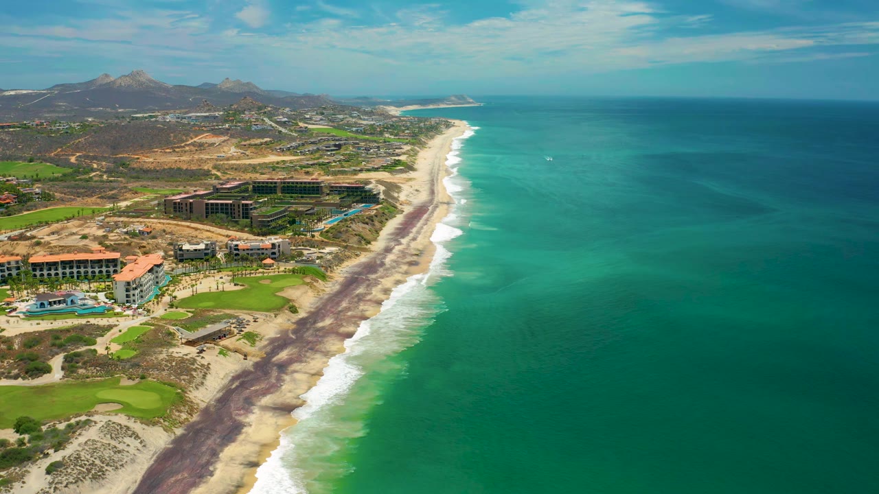 Aerial frame from San Jose del Cabo showing the beach curving into the distance, open teal ocean to the right, resort structures and fairway grass to the left, with no visible people.