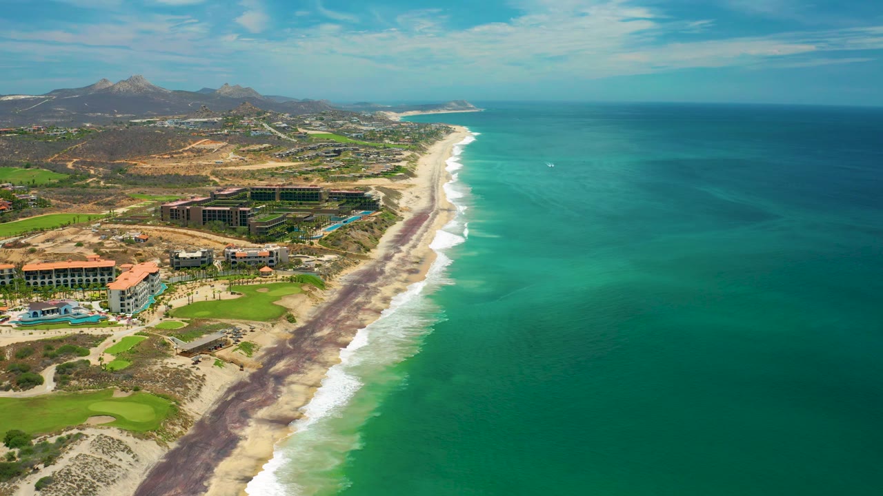 San Jose del Cabo aerial view with more open water filling the right side, a pale beach and white surf line through the center, low resort buildings and landscaped greens on the left, and no visible people.