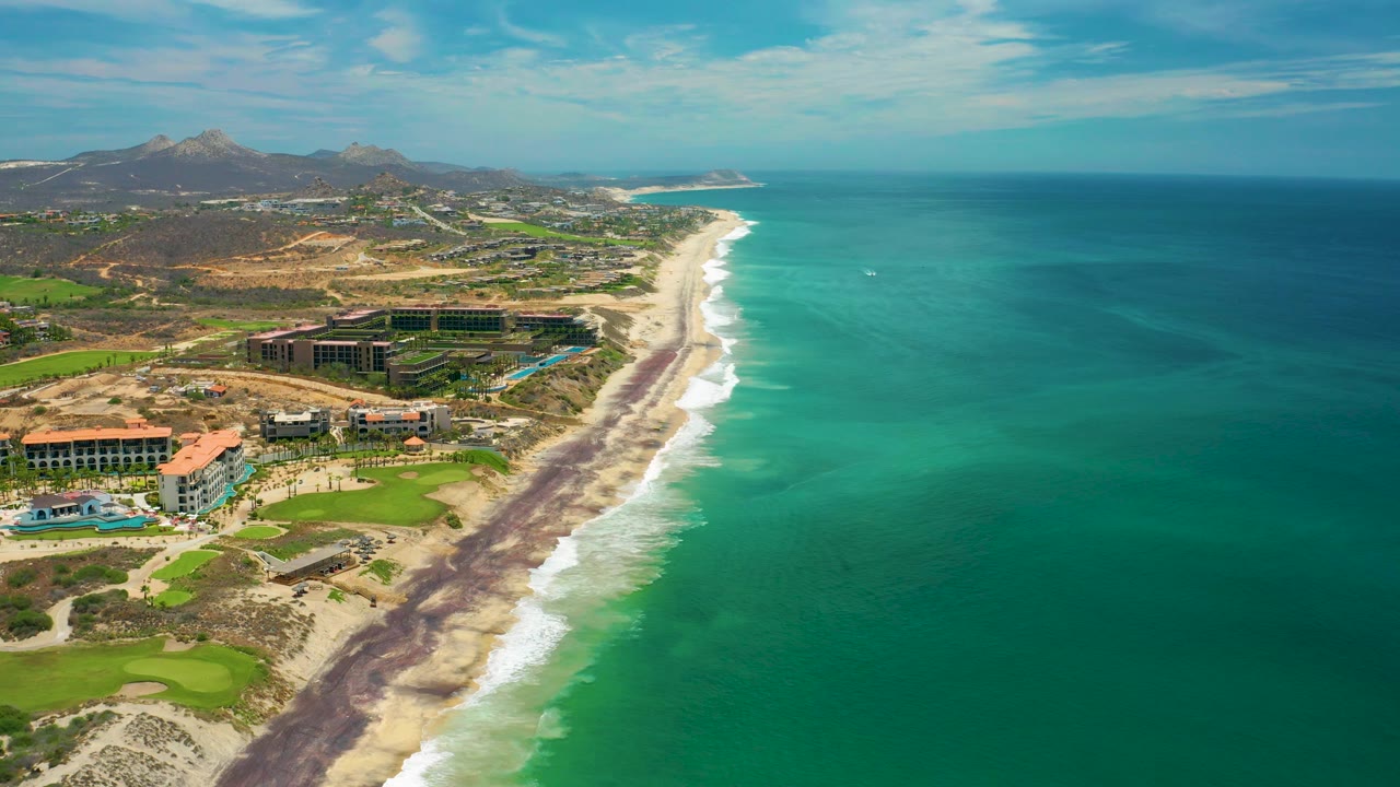 Wide aerial frame over San Jose del Cabo with the coastline running diagonally, dark material along the beach edge, bright surf, ocean to the right, resort buildings and golf greens to the left, and no visible people.