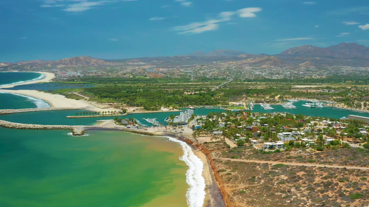 San Jose Del Cabo aerial view with a curved beach at left, sheltered turquoise water, clustered buildings, green inland areas, mountains in the distance, and no visible people.
