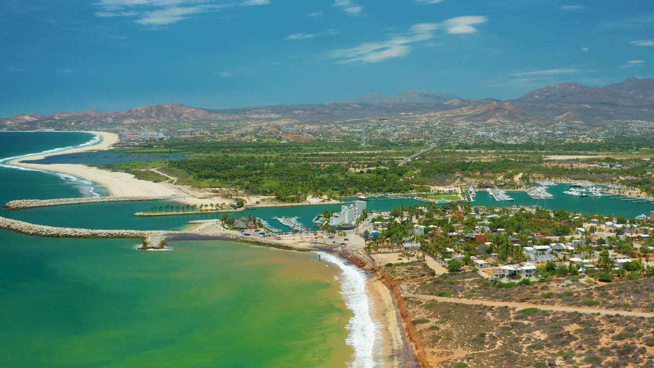 Aerial view over San Jose Del Cabo showing green-blue water, a sandy beach, marina basins, low coastal buildings, inland hills, and no visible people.