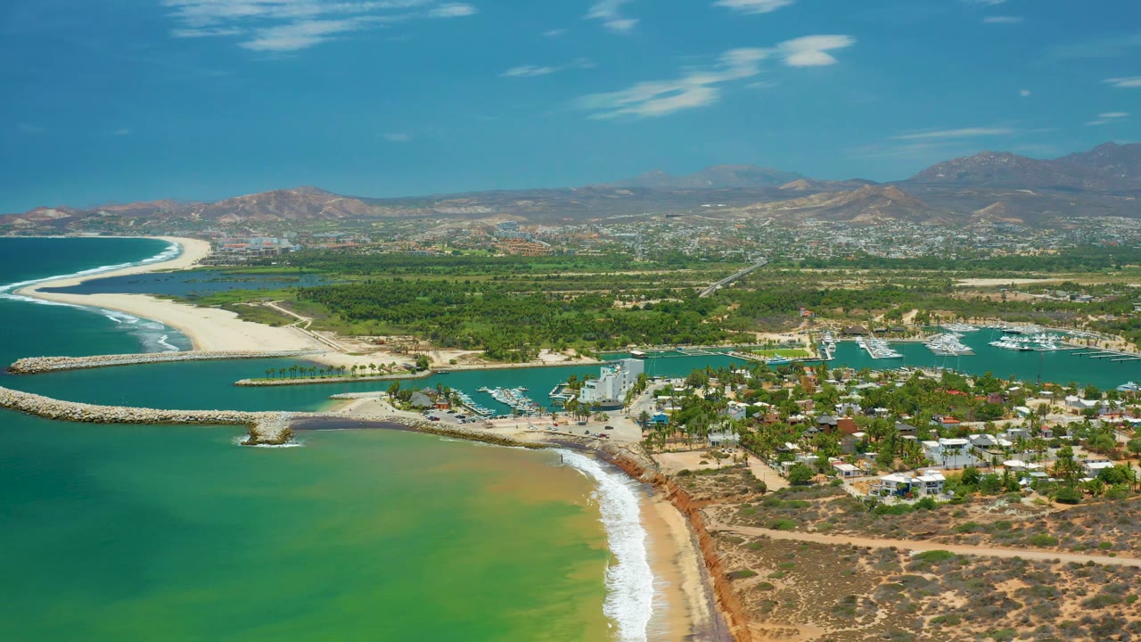 Wide aerial over San Jose Del Cabo with pale sand, a rock harbor entrance, green coastal water, a marina, clustered roofs, distant hills, and no visible people.