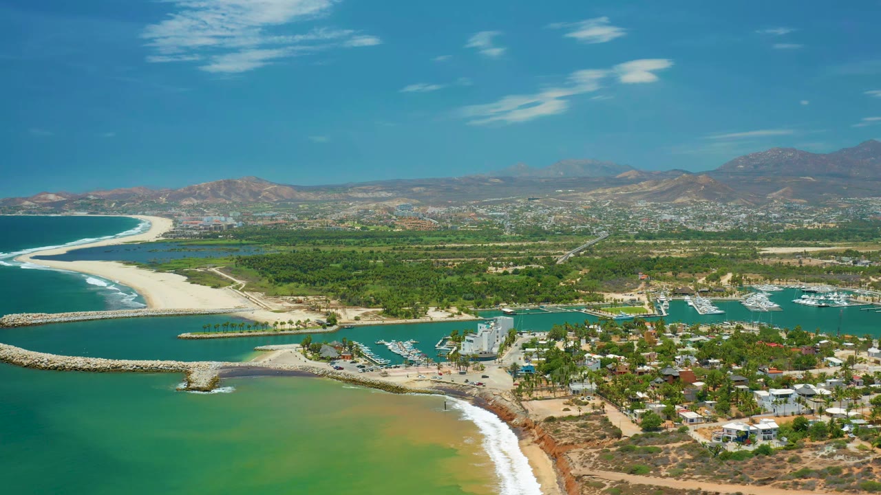 Wide aerial frame in San Jose Del Cabo showing a sandy spit at left, calm marina water, rows of docked boats, waterfront buildings, green land, mountains, and no visible people.