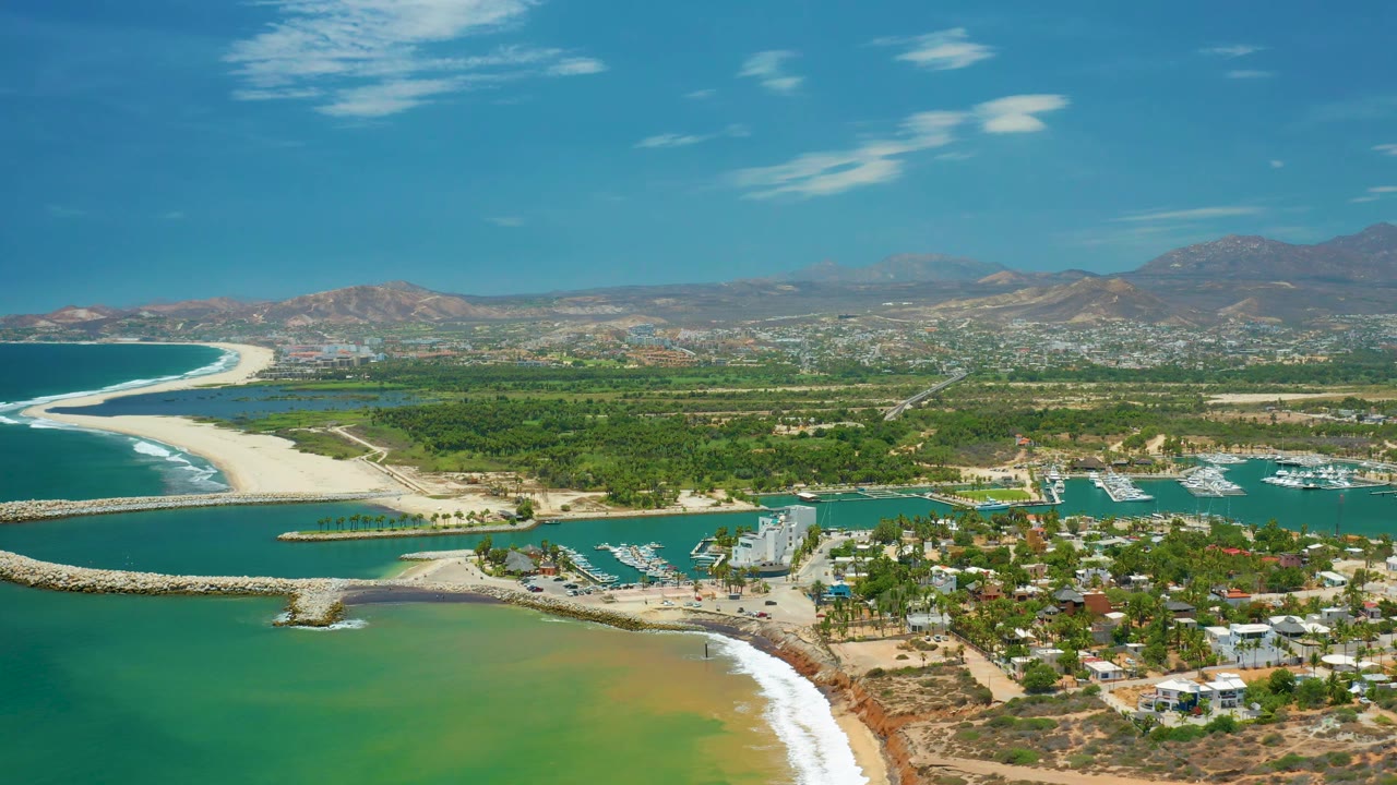 Aerial view of San Jose Del Cabo with the beach sweeping along the left side, green-blue water in front, marina docks and boats to the right, town development beyond, mountains, and no visible people.