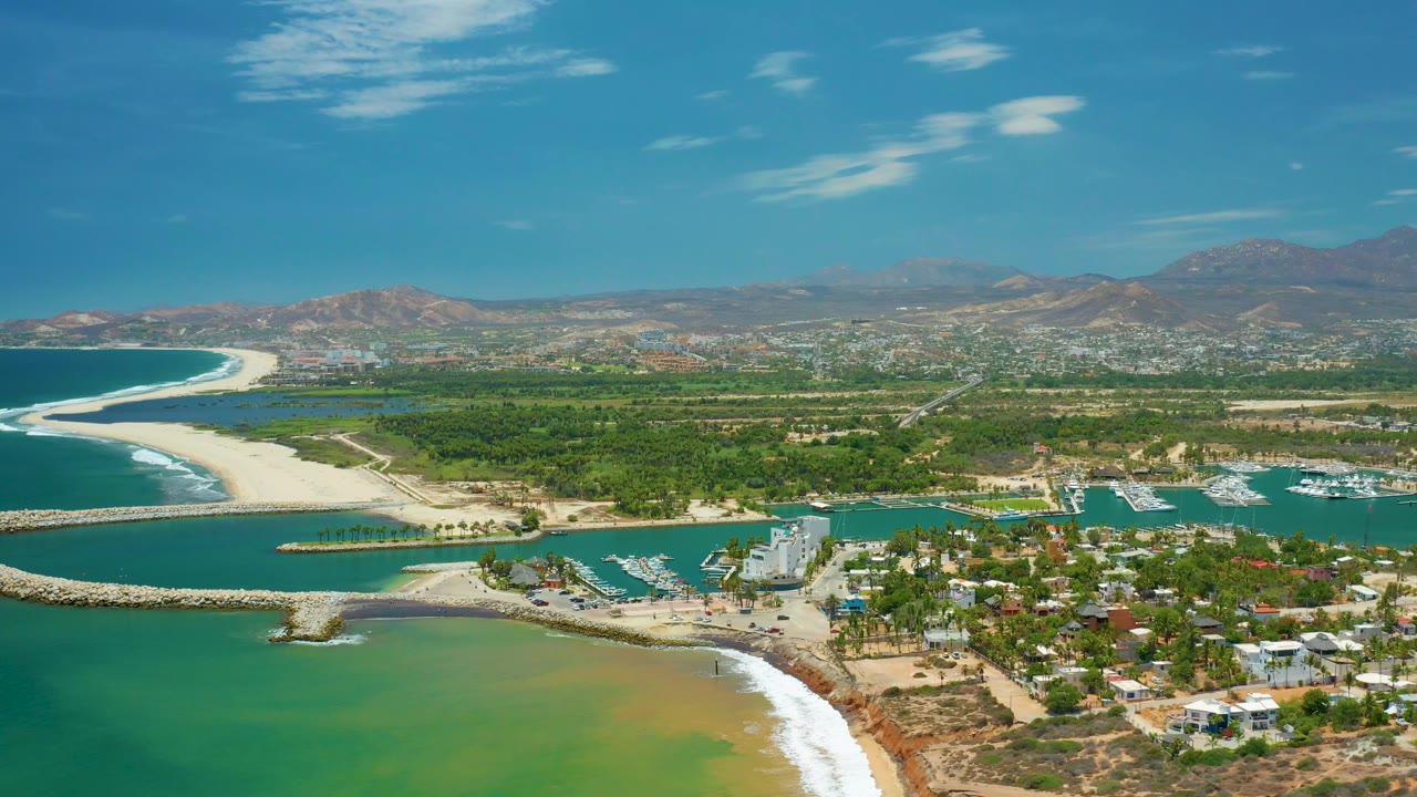 Aerial coastline with surf, breakwater, marina, buildings, and green inland area.