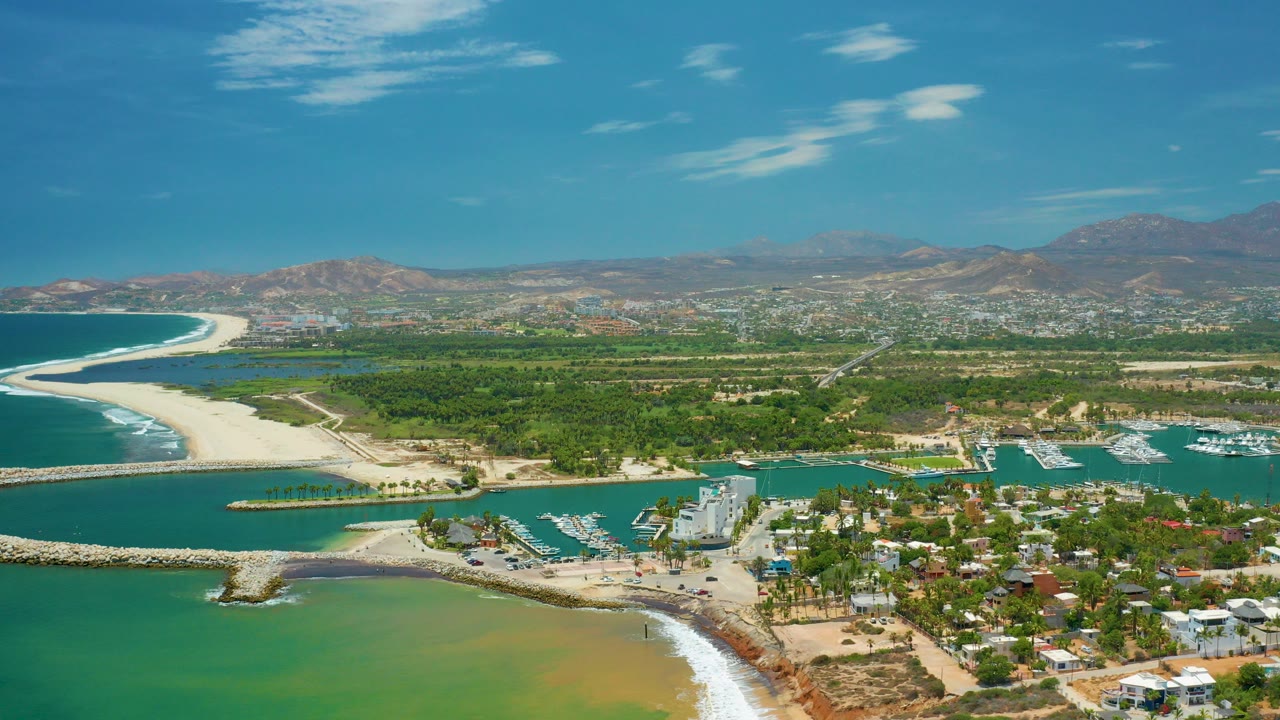 Wide aerial view over San Jose Del Cabo, Mexico, showing green-blue coastal water, sandy beach, marina slips, houses, vegetation, distant hills, and no visible people.
