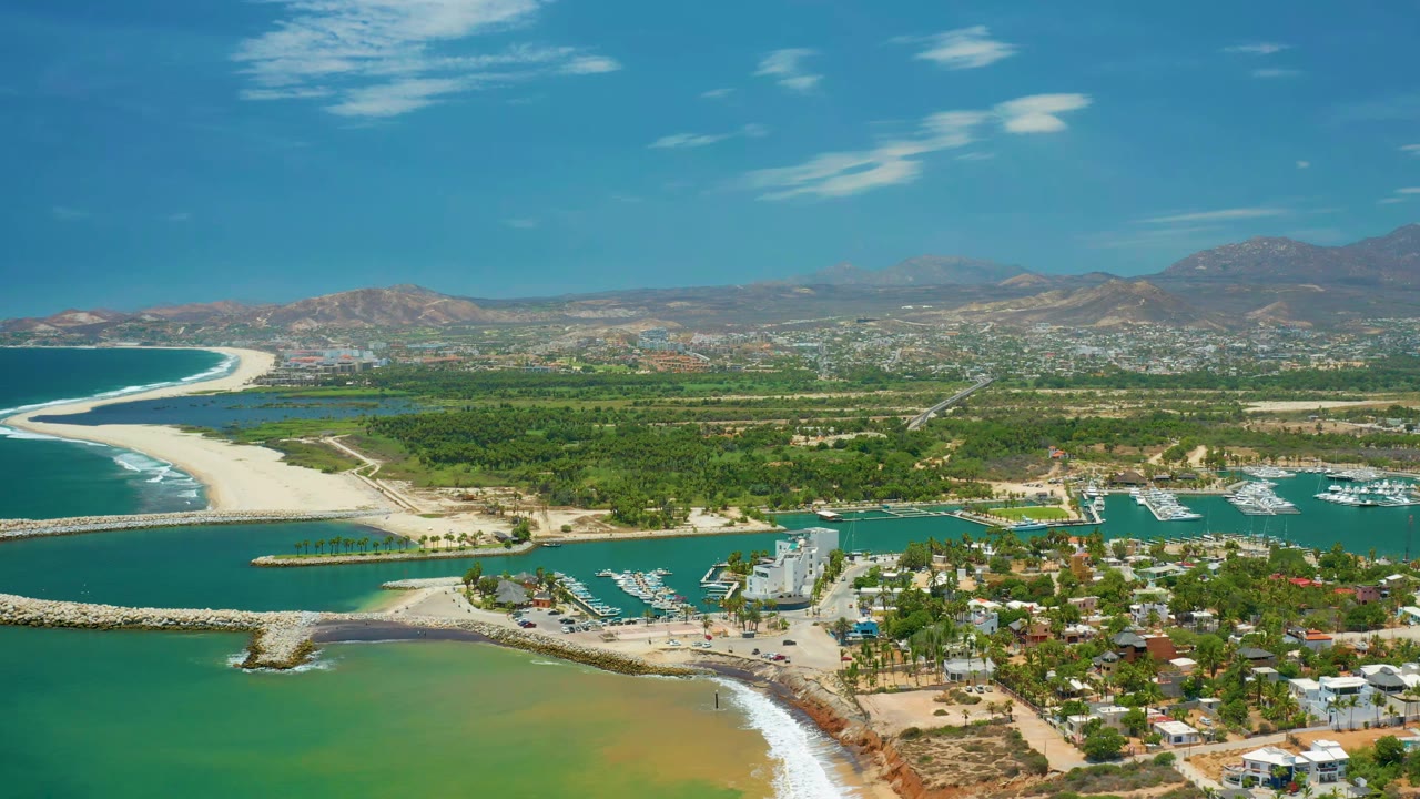 Coastal aerial with curved beach, marina water, houses, greenery, and distant hills.