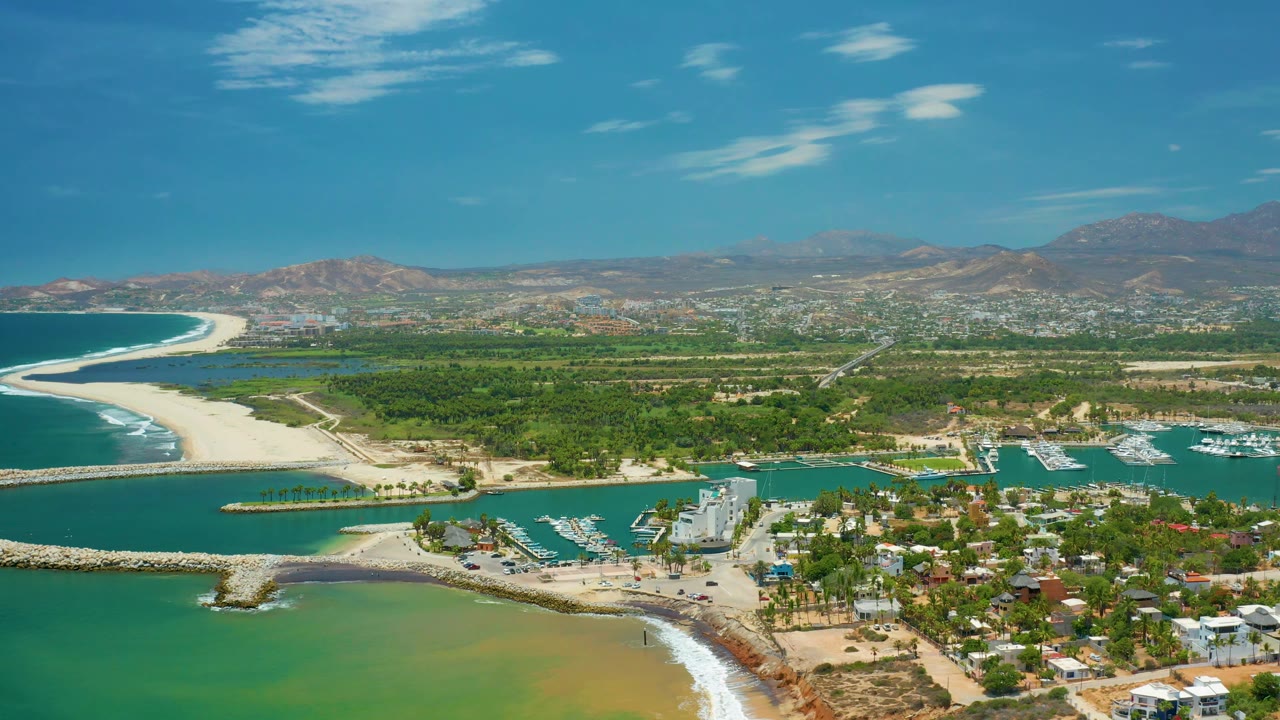 San Jose Del Cabo, Mexico appears from above with a sandy shoreline, rock breakwaters, marina docks, residential buildings, green land, hazy hills, and no visible people.