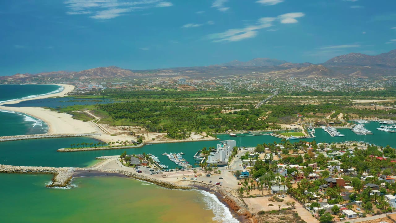 Aerial view in San Jose Del Cabo, Mexico with the curved beach and ocean on the left, marina docks near center right, low coastal buildings in front, distant mountains, and no visible people.