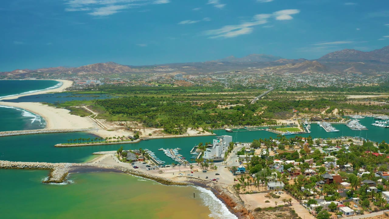 San Jose Del Cabo aerial frame showing the white sand spit at left, marina boats and docks on the right side of the waterway, a cluster of low buildings below, mountains beyond town, and no visible people.
