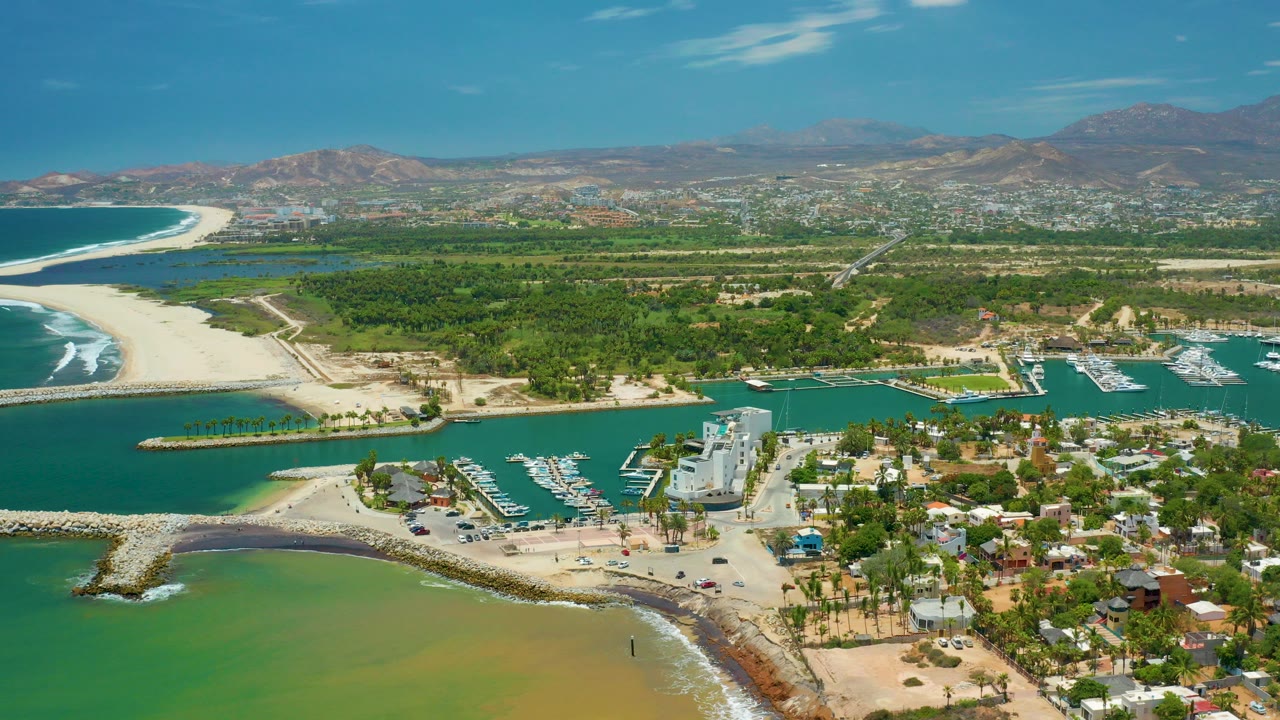 Opening aerial frame over San Jose Del Cabo, Mexico, showing turquoise inlet water, sandy beaches, marina docks, coastal buildings, green vegetation, distant mountains, and no visible people.