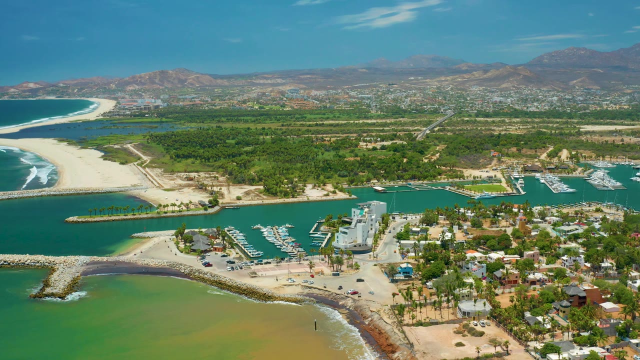 Aerial view in San Jose Del Cabo, Mexico, showing turquoise marina water across the lower frame, sandy shorelines, clustered coastal buildings, green land, hazy mountains, and no visible people.