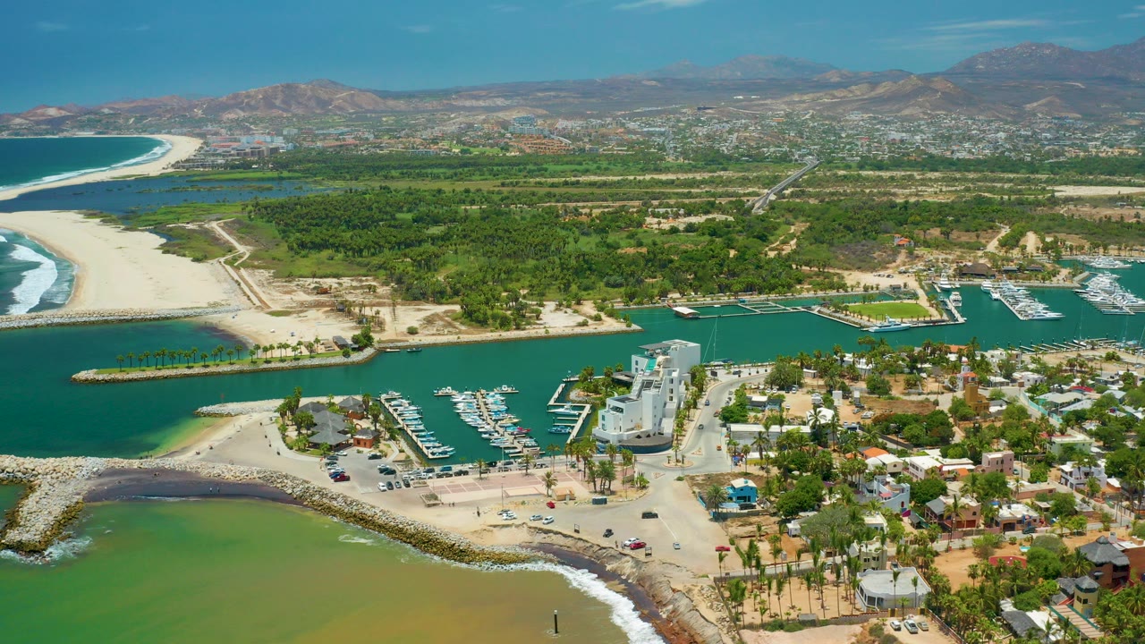 Wide aerial frame of San Jose Del Cabo showing the inland marina, sandy inlet, green coastal flats, town buildings, distant hills, and no visible people.