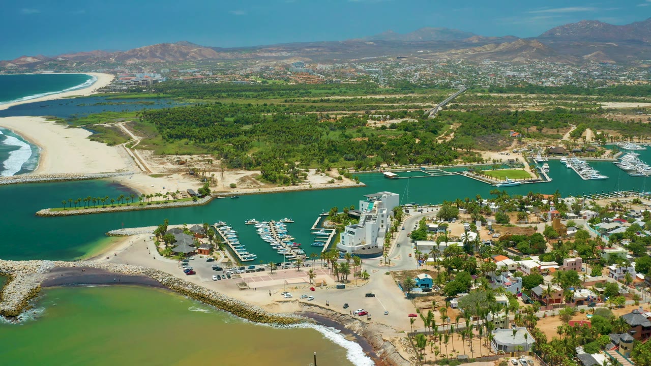 High aerial frame of San Jose Del Cabo with the marina entrance, rows of boats, surf wrapping the sandy inlet, roads, palms, inland greenery, and no visible people.
