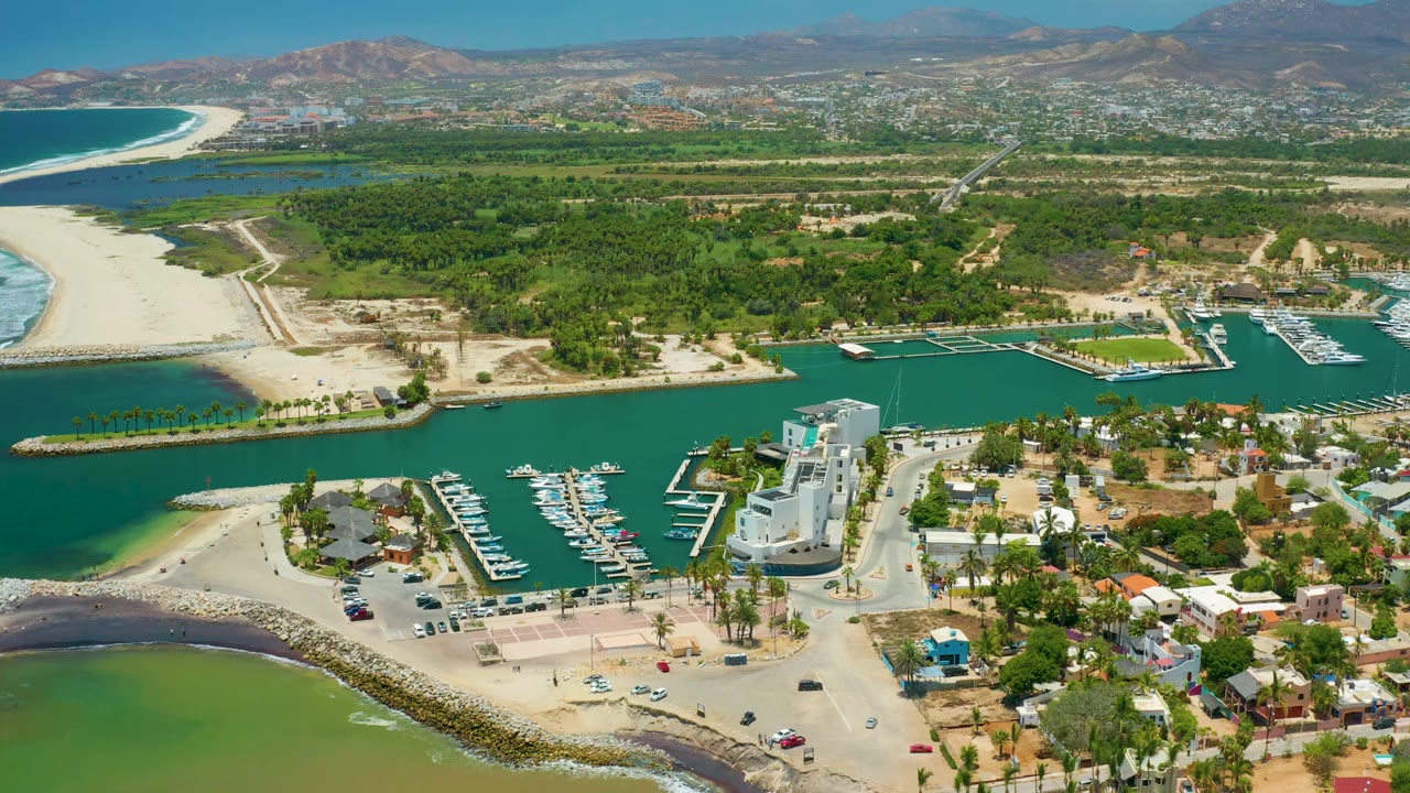 Aerial frame over San Jose Del Cabo with docked boats in the marina foreground, a pale beach at left, roads and buildings below, and no visible people.