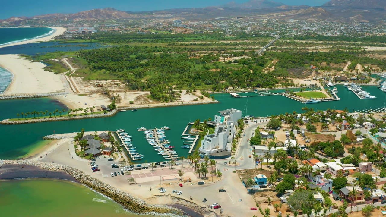 San Jose Del Cabo aerial view showing turquoise port water, clustered docks, shoreline buildings, a green coastal band, distant urban area, and no visible people.