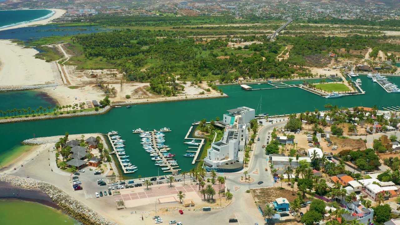 Second aerial frame of San Jose Del Cabo, Mexico with rows of boats in calm harbor water, a white building cluster near the road, sandy beach edges, and dense green land beyond, with no visible people.