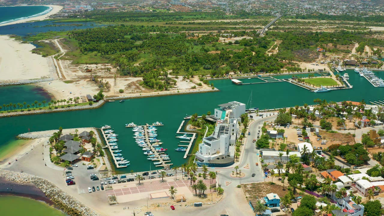 Aerial view in San Jose Del Cabo, Mexico showing the marina basin, several docks with boats, white shore buildings, palm-lined streets, sandy beach, and green vegetation in the distance, with no visible people.
