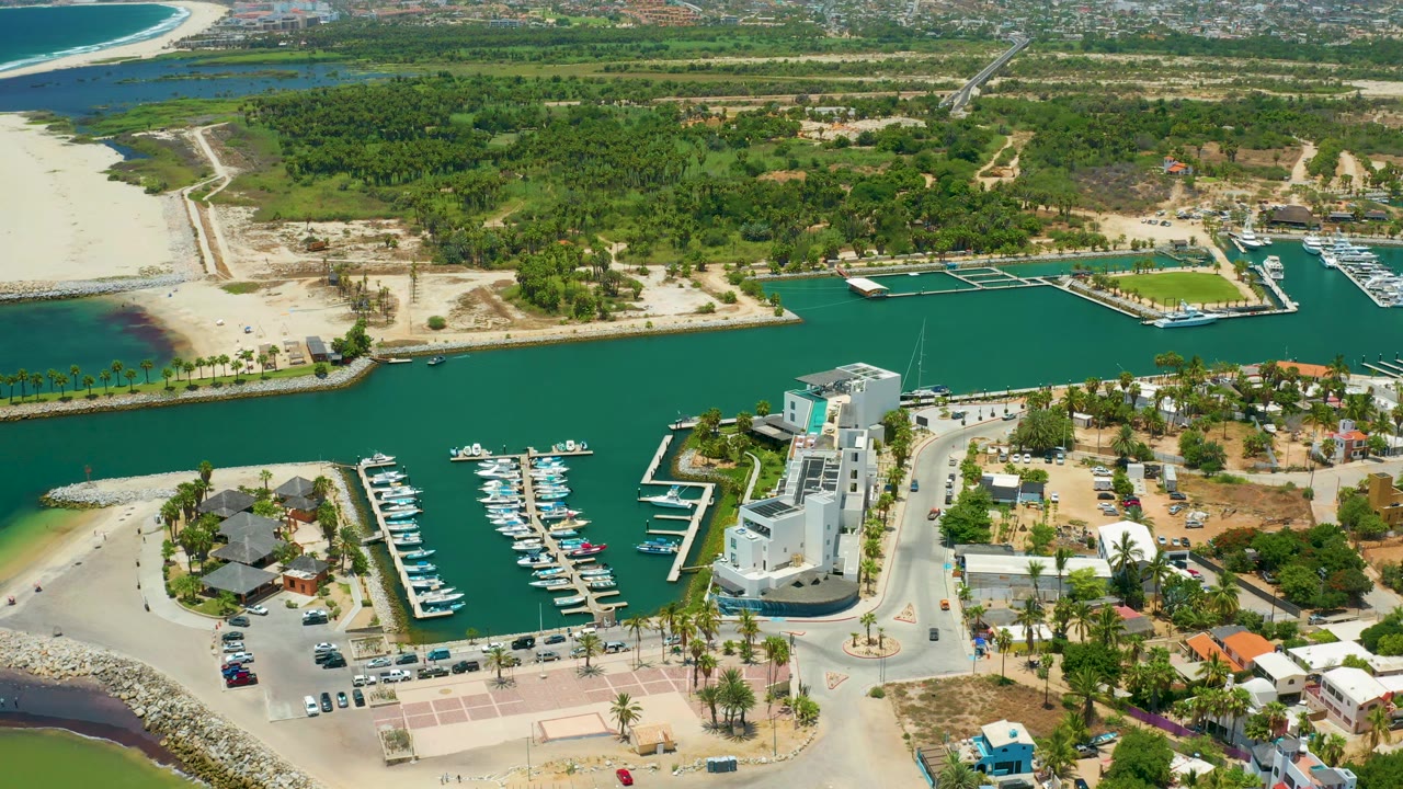 Wide aerial view of turquoise harbor water, docked boats, white buildings, sandy shoreline, and green trees.