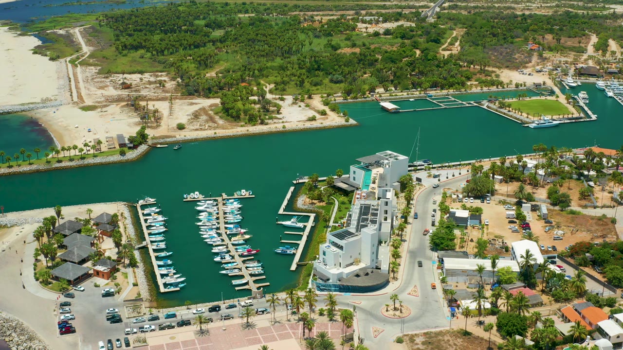 Aerial marina view with boats in slips, a white building, waterfront road, beach, and inland greenery.