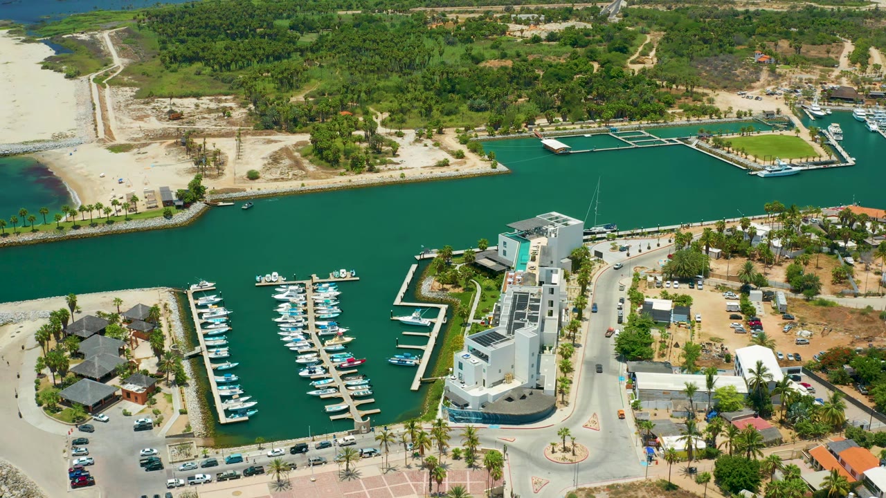 Aerial view in San Jose Del Cabo, Mexico showing the white waterfront structure closer to the lower center, marina slips to both sides, cars along the road, and a sandy beach crossing the upper left.