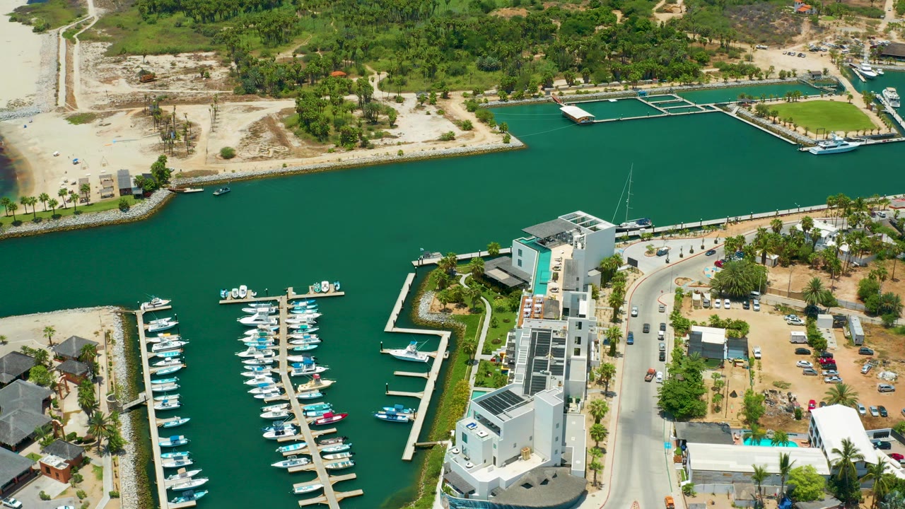 Aerial view over San Jose Del Cabo harbor with green water, docked boats at white piers, a curved road, waterfront buildings, sandy shoreline, and no visible people.