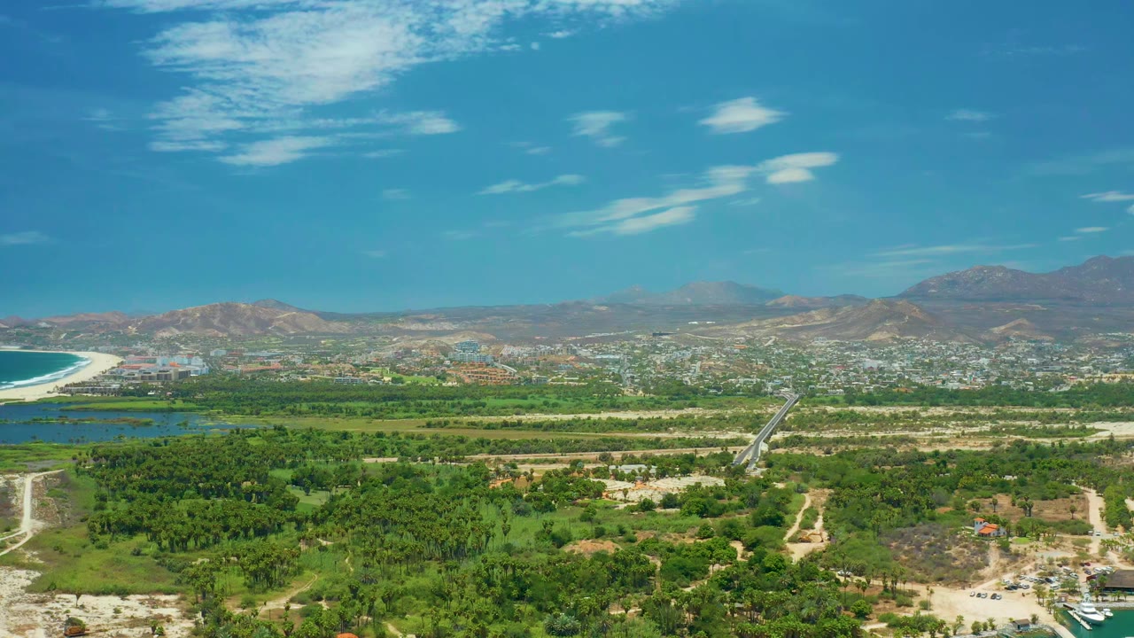 Aerial view in San Jose del Cabo, Mexico, with the coastline sitting at the left edge, dense green lowland below, town structures in the distance, hills on the horizon, and no visible people.