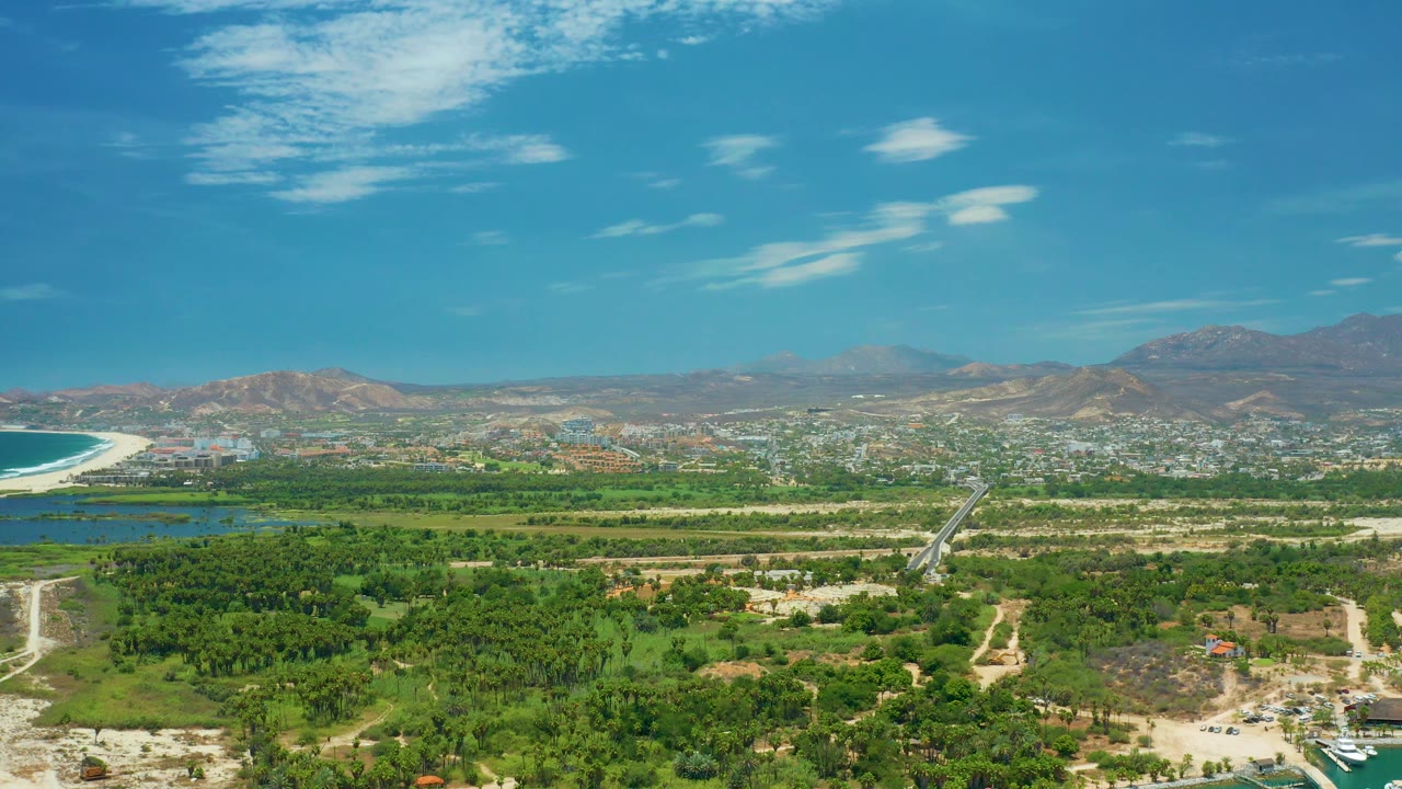 High aerial frame over San Jose del Cabo, Mexico, showing a pale road cutting through green land toward distant buildings, ocean on the left, brown hills behind the town, and no visible people.