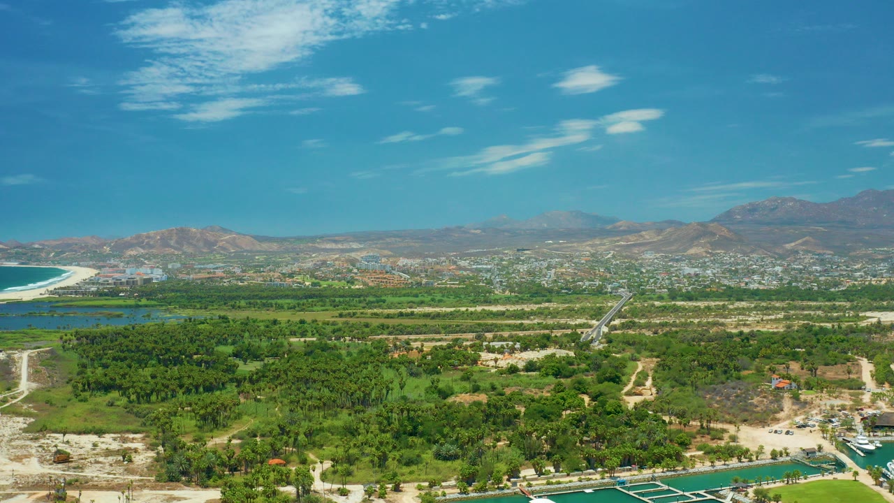 Opening wide aerial frame over San Jose del Cabo with marina water at the bottom, dense green vegetation, distant town, dry mountains, scattered clouds, and no visible people.