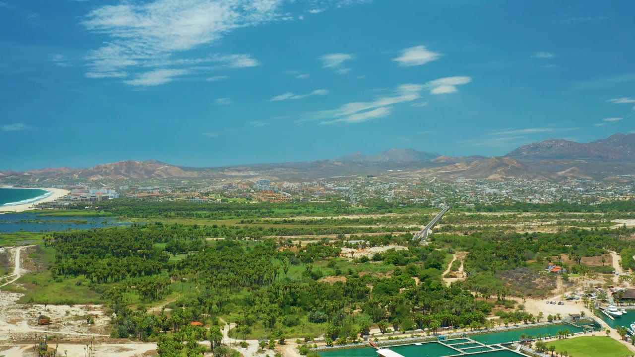 Third frame shows San Jose del Cabo from above with a stronger view of the winding road through green trees, water along the lower edge, distant town blocks, low mountains, and no visible people.