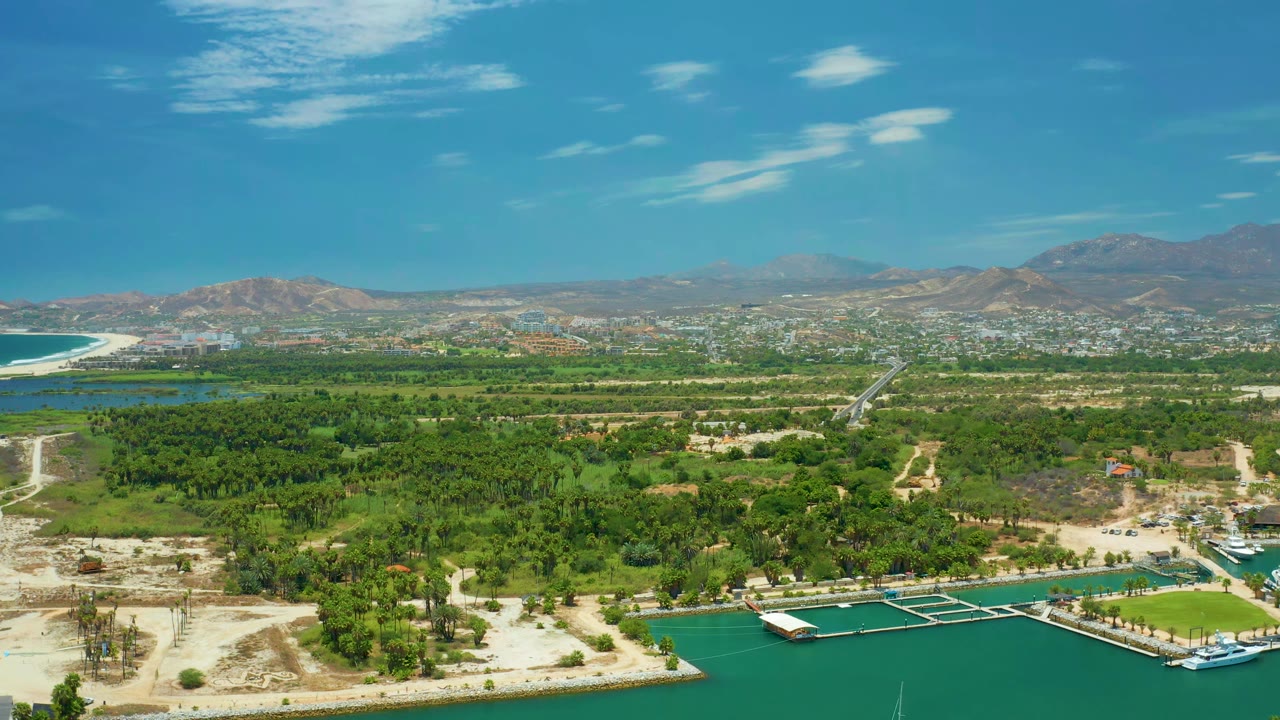 An elevated San Jose del Cabo view holds on the marina edge, with dock slips and small boats below, a green peninsula through the middle, city blocks in the distance, and no visible people.