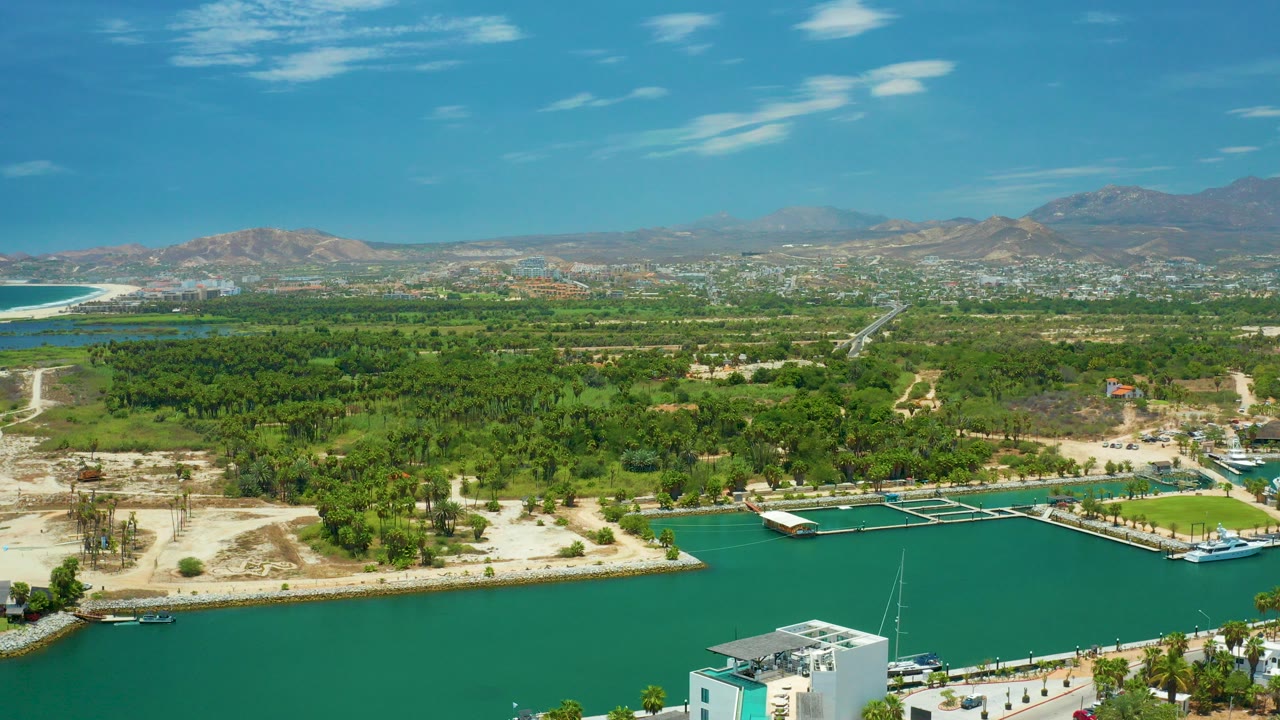 Aerial view in San Jose Del Cabo, Mexico, looking over turquoise marina water, docks, green trees, palms, distant buildings, and brown mountains under blue sky, with no visible people.