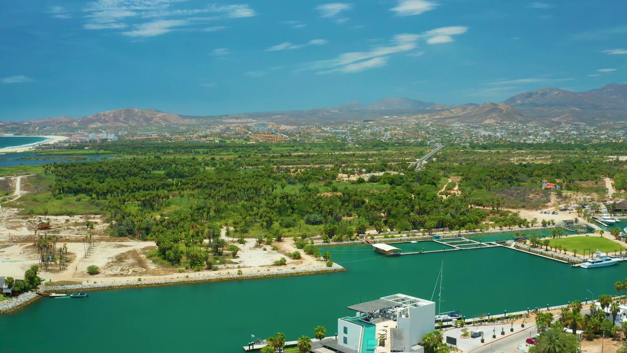 Aerial view in San Jose Del Cabo with a large area of turquoise water, small white boats near marina slips, dense green shoreline vegetation, pale sandy ground, and dry mountains in the distance, with no visible people.