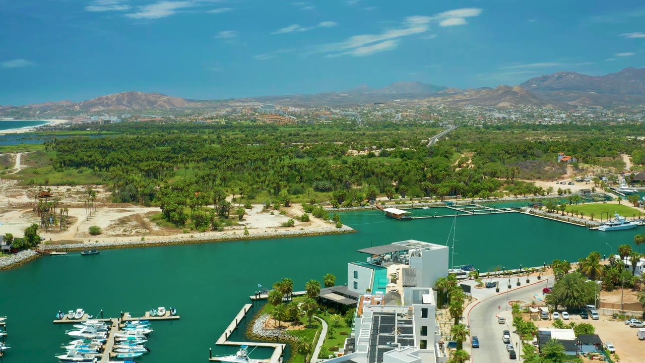 At 0:01, the aerial view in San Jose Del Cabo holds on the marina basin, with boats at the lower left, a white building near the shoreline, dry lots, green tree cover, mountains, and no visible people.