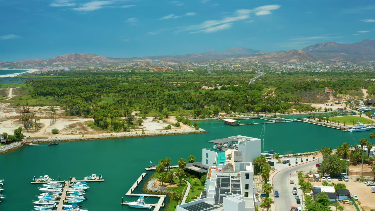 At 0:02, San Jose Del Cabo appears from above with the turquoise channel across the foreground, boats clustered at lower left, a palm-lined curve of road, low buildings, green inland terrain, and no visible people.