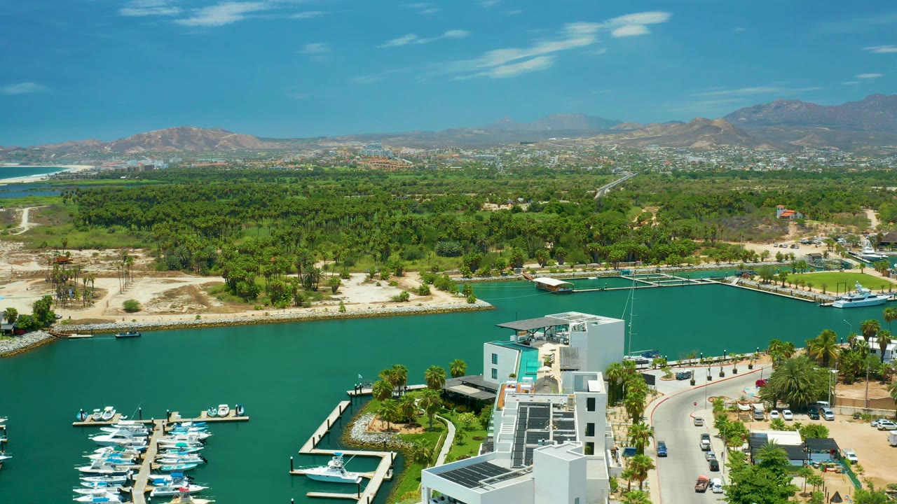 At 0:03, the view looks across San Jose Del Cabo marina water toward a green shoreline, dock structures, sandy open areas, distant buildings at the base of the mountains, and no visible people.