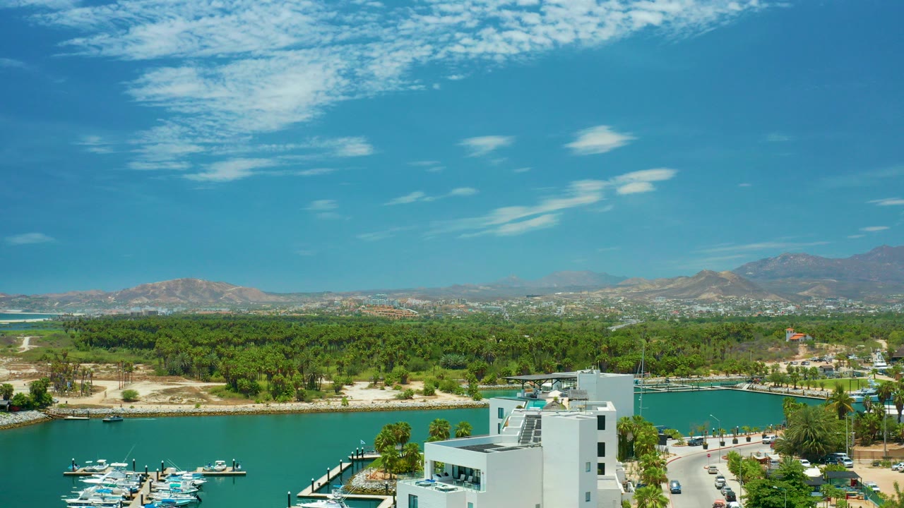 Aerial view of turquoise marina water, white buildings, trees, mountains, and blue sky.