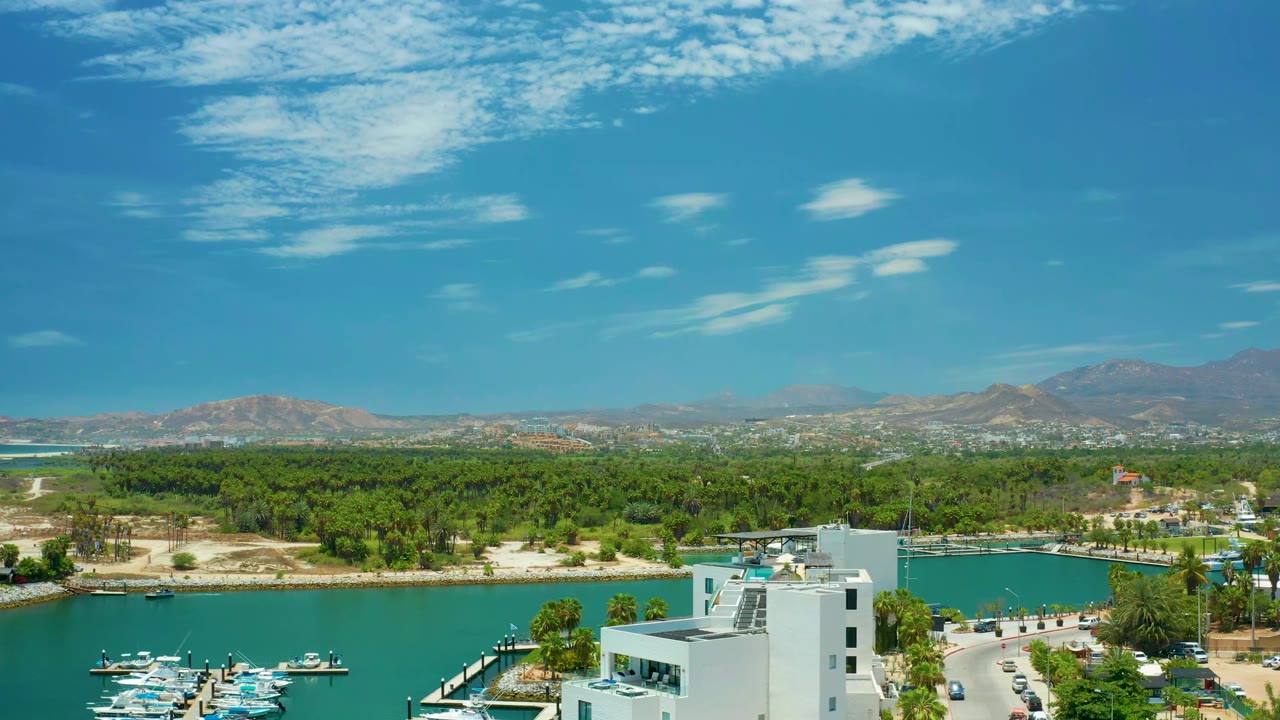 At 0:01, the San Jose Del Cabo view holds on a white waterfront building, small boats in turquoise water, a green tree belt, low mountains on the horizon, thin clouds, and no visible people.