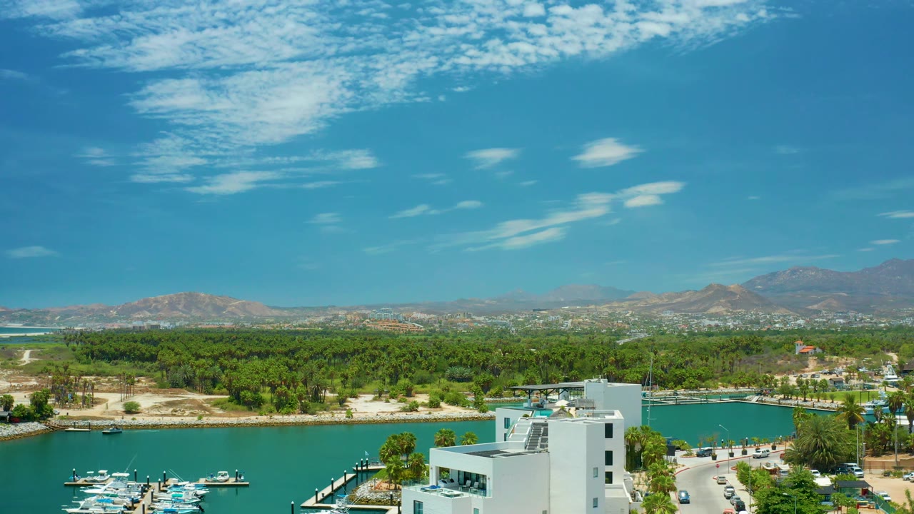 At 0:02, the elevated San Jose Del Cabo composition shows boats clustered near docks, a white building at lower right, green shoreline trees, hazy mountains, textured cloud patches, and no visible people.