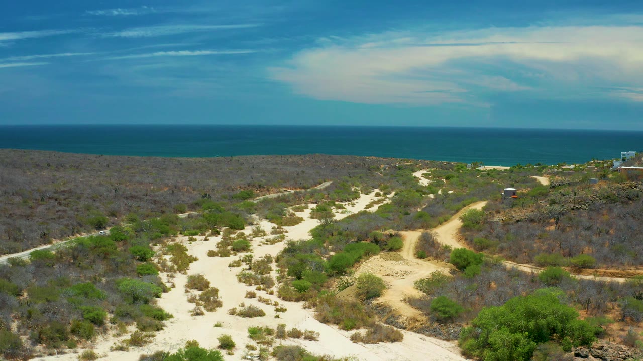 A wide elevated frame over San Jose del Cabo shows pale sand paths, low bushes, a small distant structure near the right edge, blue water beyond the land, and no visible people.