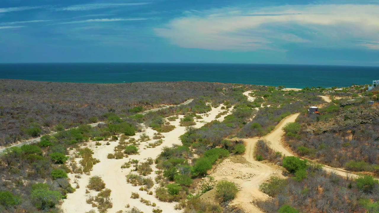 Sandy washes and dirt paths run through scrubland toward the blue ocean horizon in San Jose Del Cabo, Mexico, with no visible people.