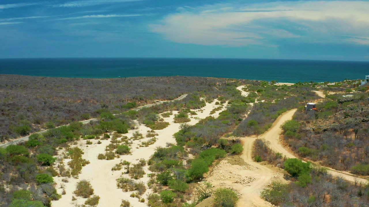 Wide aerial desert view with sand, scrub, and ocean beyond.