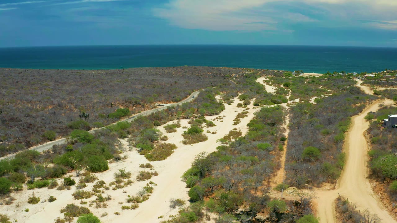 Elevated San Jose del Cabo view with a broad pale sand area in the foreground, scattered green shrubs, gray brushland, a tan track at right, ocean beyond, and no visible people.
