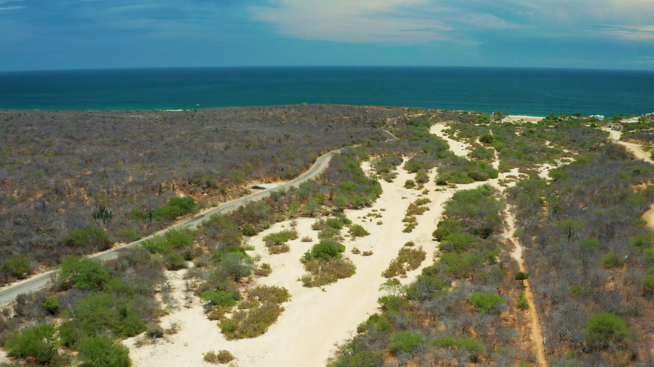 Opening aerial view in San Jose del Cabo, Mexico, showing pale sandy trails, scrub vegetation, a narrow road at left, blue sea and sky in the background, and no visible people.