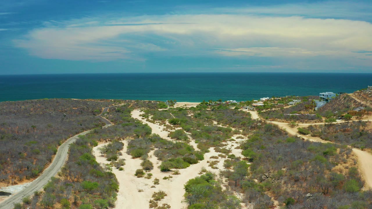 Aerial frame from San Jose Del Cabo, Mexico showing pale sand paths through low coastal vegetation, a curving road at left, turquoise sea in the distance, and no visible people.