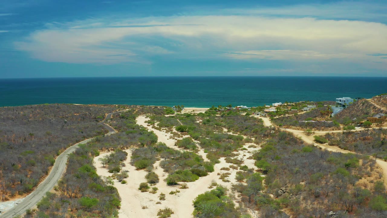 San Jose Del Cabo aerial frame showing a road bending through dry scrubland, pale sand channels down the center, a narrow beach, distant homes, and no visible people.