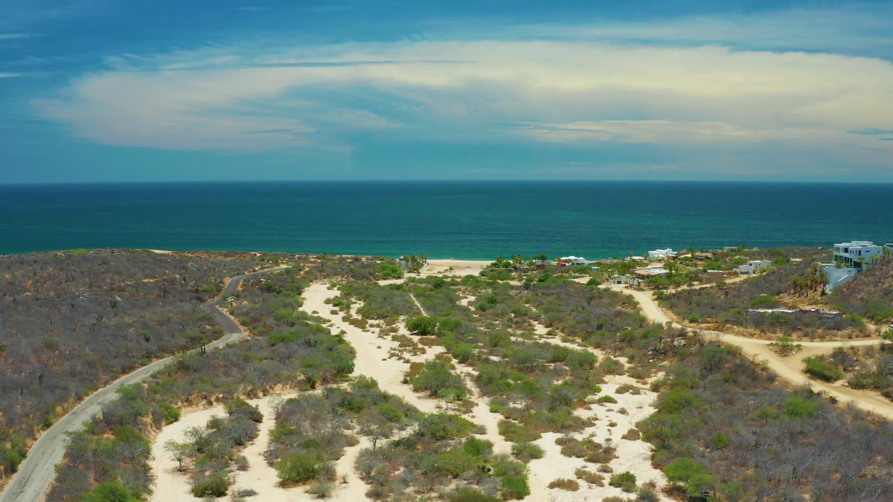 Opening frame of San Jose Del Cabo coastline with pale sandy tracks through scrubland, the beach and blue sea beyond, a large cloud band overhead, and no visible people.