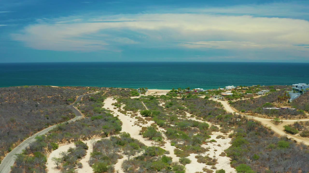 San Jose Del Cabo coastline seen from above with dry scrubland in the foreground, branching sandy paths, a narrow beach strip, open blue water, and no visible people.