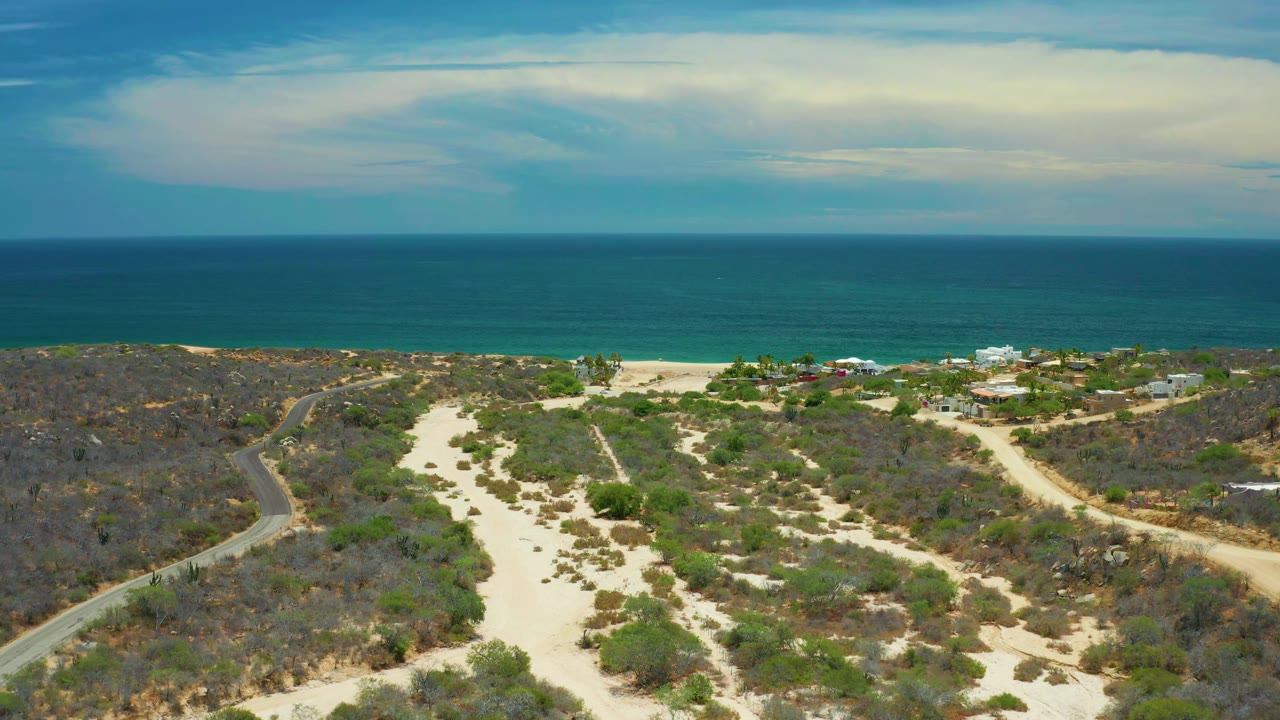 Aerial coastline view with sand tracks, scrub, beach buildings, and ocean horizon.